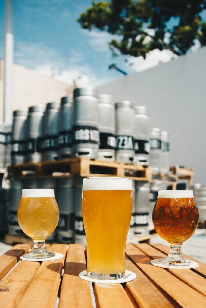 pexels-photo-1267700-1267700 Assorted beer glasses on a wooden table with brewery kegs in the background.