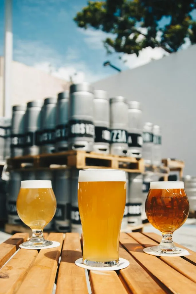 pexels-photo-1267700-1267700 Assorted beer glasses on a wooden table with brewery kegs in the background.